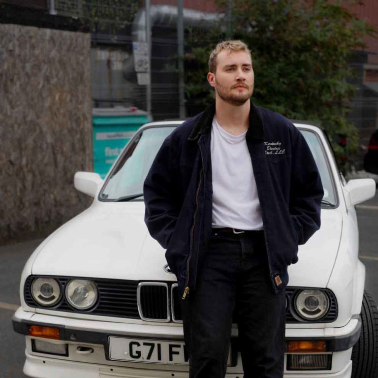 A portrait of Sam Wills leaning against a white BMW.